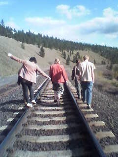 Grainy flip phone image shows a group of young people walking away from the camera on train tracks. One is balanced on the rail with their arms splayed to the sides. The sky is blue and partly cloudy.