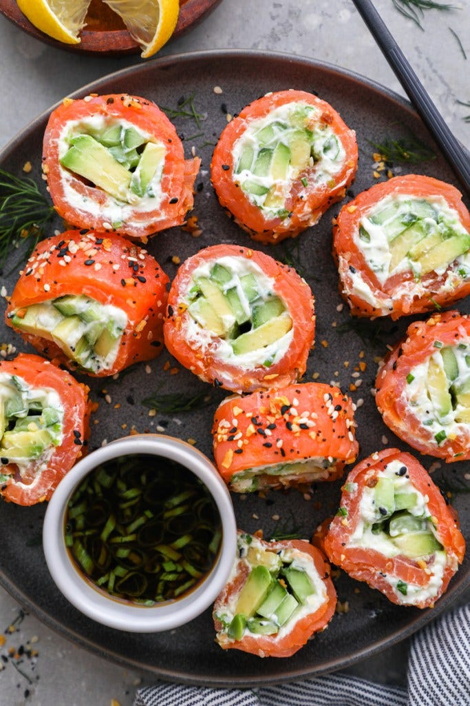 Smoked salmon roll ups sitting on a dark plate next to a small bowl of citrus soy sauce