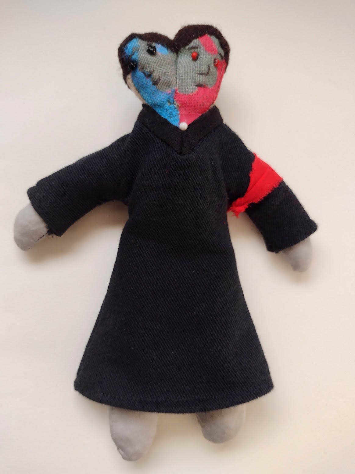 Fabric doll with two heads- one with red streaks and one with blue, wearing a black judge's robe and a red armband