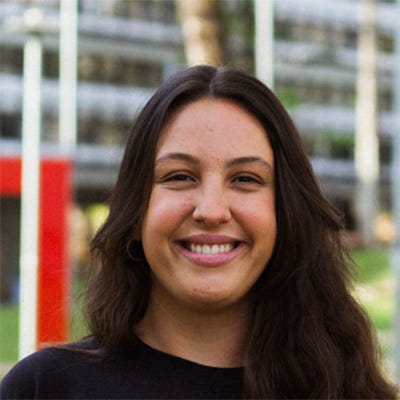 Mulher de camiseta preta com cabelos escuros e compridos, sorrindo, tendo ao fundo um edifício com gramado onde está instalada uma escultura Mulher de camiseta preta com cabelos escuros e compridos, sorrindo, tendo ao fundo um edifício com gramado onde está instalada uma escultura