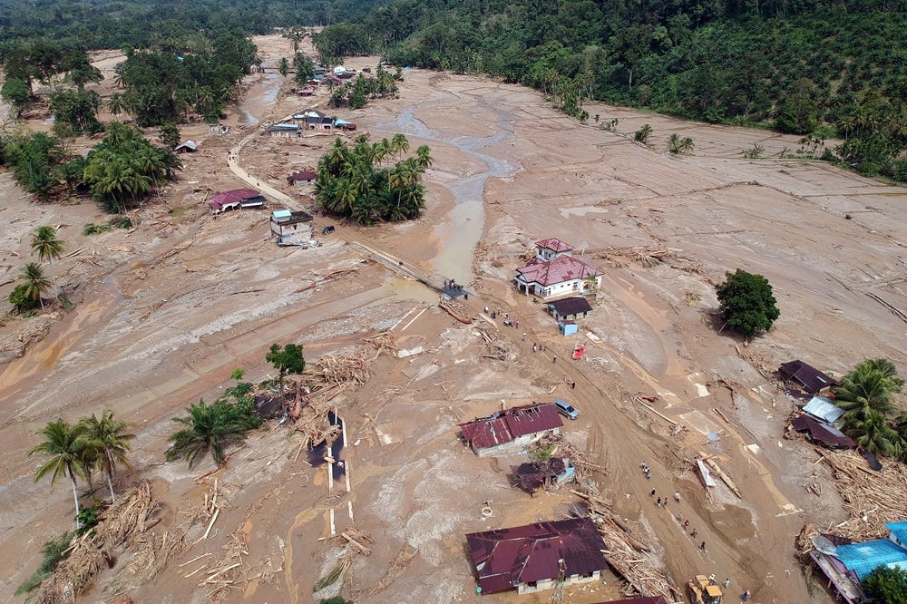 Mud and brown water flooded a rural area amidst green forests in a valley, with some roofs of houses visible sticking out in a state of severe damage.