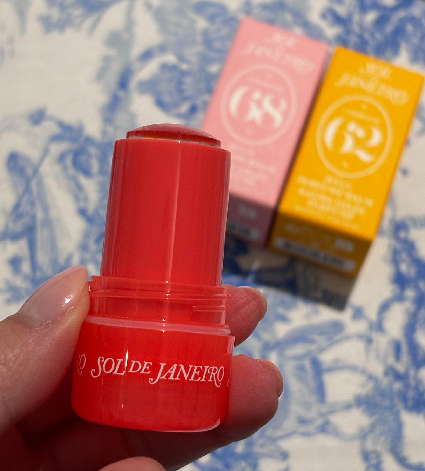 A close-up view of a hand with light fingernail polish holding an open bright pinkish-red Sol de Janeiro stick applicator. The balm product is twisted up and visible. In the background, out of focus, are the product boxes for '68' (pink) and '62' (yellow).