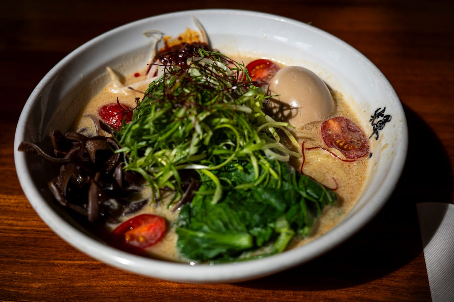 A bowl of ramen soup with shredded mushrooms, cherry tomatoes, spinach, and a pickled egg.