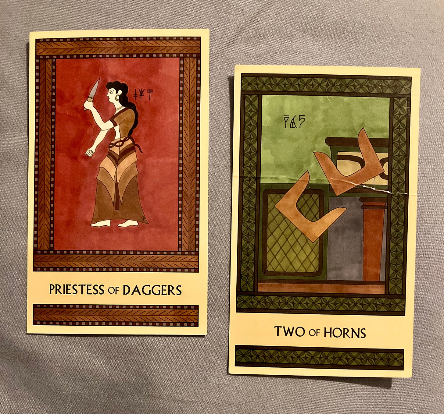 Two Minoan Tarot cards on a gray background. ThePriestess of Daggers is painted in shades of deep red and brown. It shows a Minoan woman standing, facing left, brandishing a dagger. The Two of Horns is painted in shades of earthy green and brown. It shows two pairs of Minoan sacred horns in midair as they tumble off a shrine.
