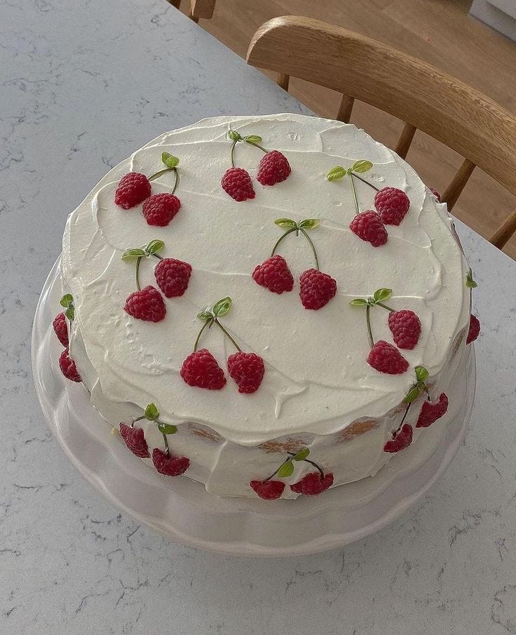 This may contain: a white cake topped with raspberries on top of a glass plate next to a wooden chair