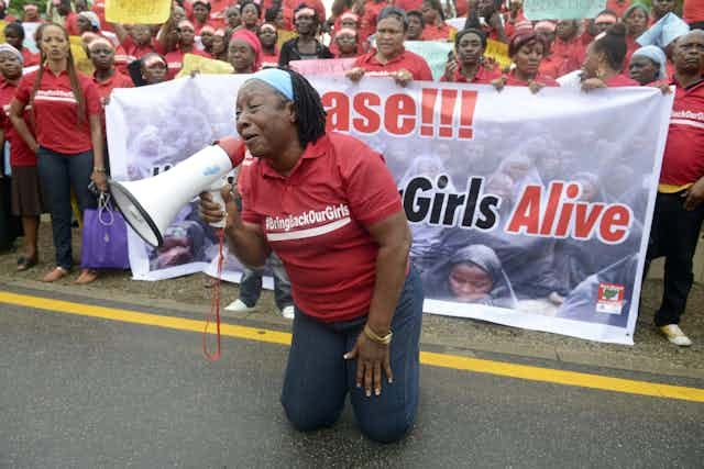 A woman in a red shirt kneels in front of a group of protestors, using a megaphone A woman in a red shirt kneels in front of a group of protestors, using a megaphone