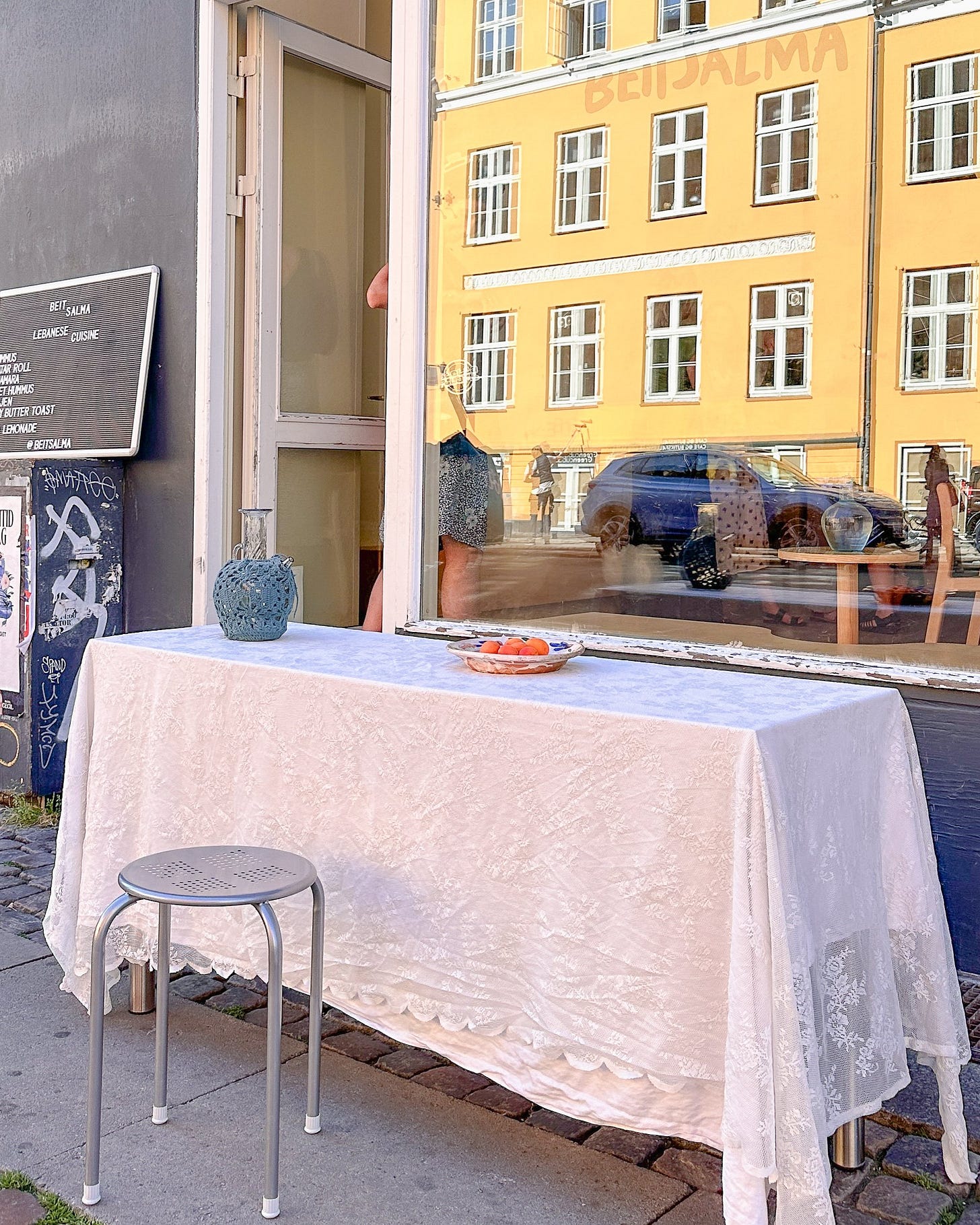 Exterior of BeitSalma, a Lebanese café in Nørrebro, Copenhagen, featuring a table with a white lace tablecloth. Discover Lebanese café culture with Scandinavian design at BeitSalma.