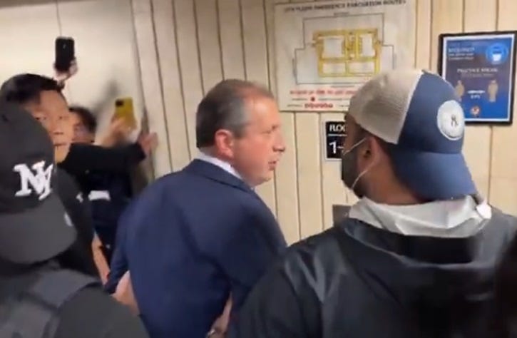 Video screenshot of Brad Lander, a balding middle-aged white man in a blue suit, being manhandled by several plainclothes ICE agents in a hallway in front of a bank of elevators.. Two of the agents wear backwards baseball caps, one wears a disposable mask. A crowd of reporters can be seen recording the arrest with their phones. Video screenshot of Brad Lander, a balding middle-aged white man in a blue suit, being manhandled by several plainclothes ICE agents in a hallway in front of a bank of elevators.. Two of the agents wear backwards baseball caps, one wears a disposable mask. A crowd of reporters can be seen recording the arrest with their phones.