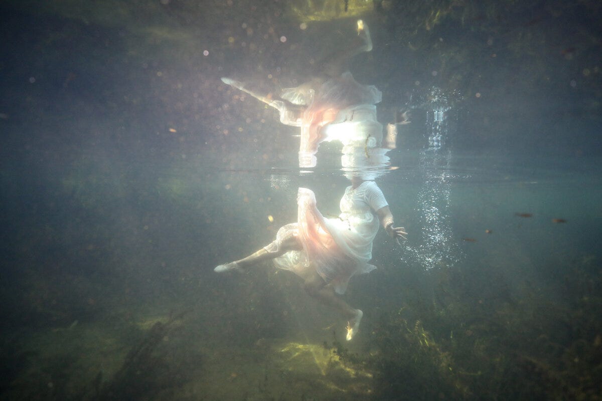 A woman in a billowing white dress is swimming in a river. The framing of this photo is below the waterline so we see her body, arms, but her head is not visible.