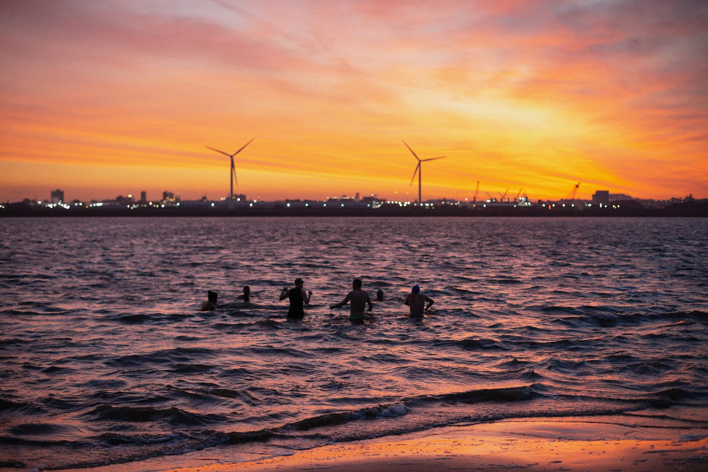 A group of people standing in the River Mersey as a strong sunrise illuminates the morning sky.
