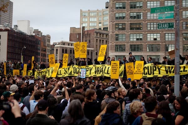A dense crowd fills a city street, many holding yellow protest signs and a banner reading "Occupy Wall Street.”
