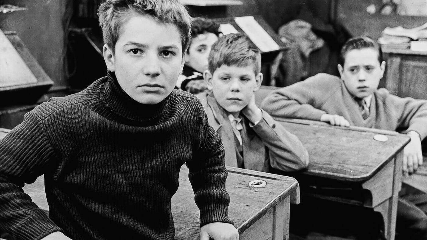 Antoine Doinel sits at his desk in a classroom, staring intently at the camera while his classmates look on behind him.