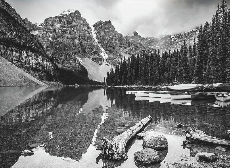 Monochrome photograph of Moraine Lake in the Canadian Rockies with still water reflecting jagged peaks and forest.