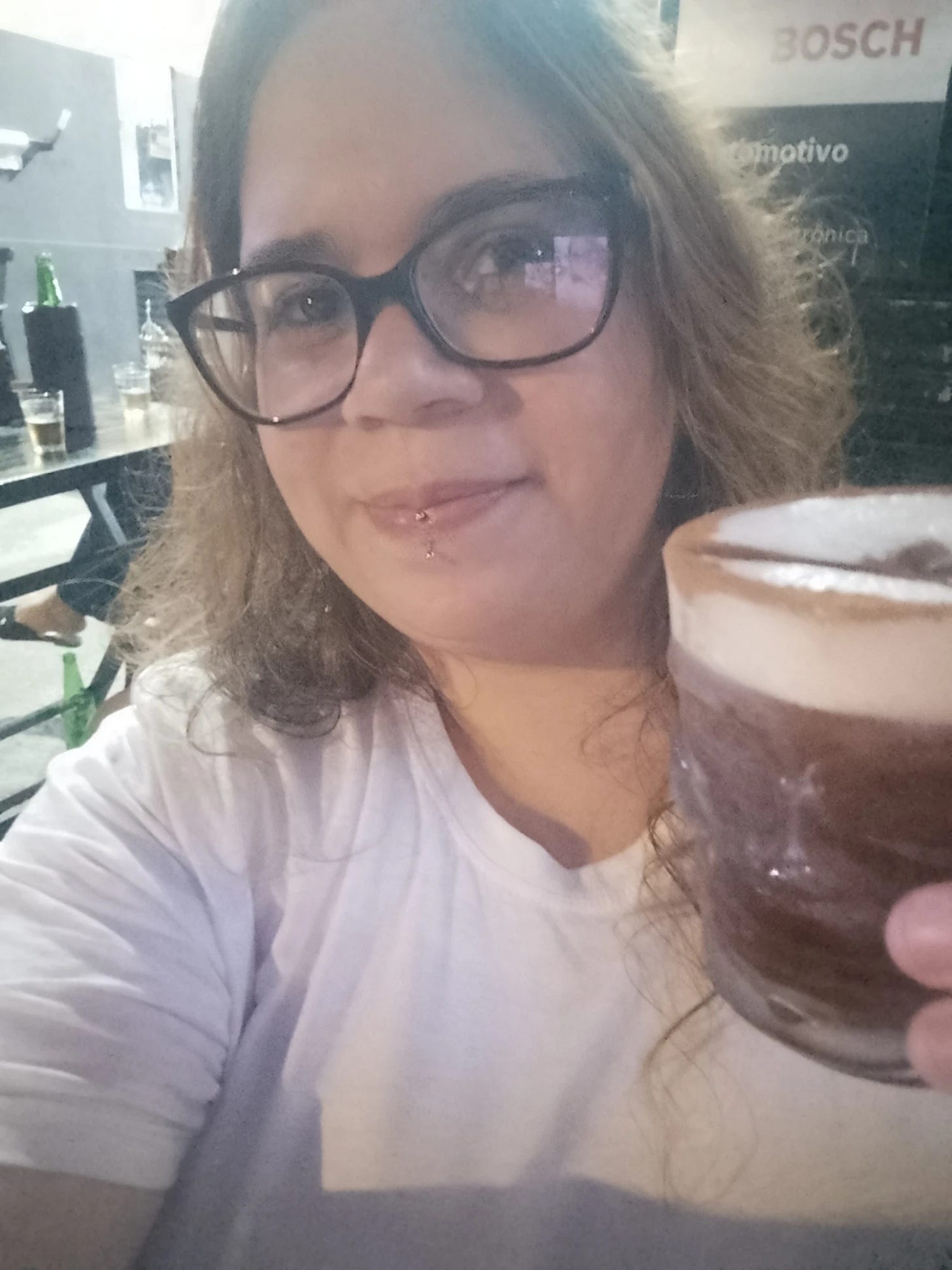 Mulher com cabelos longos e castanhos, rosto redondo, óculos grossos com armação preta, camiseta branca e piercing vertical labret. Segurando copo em formato de caveira com bebida escura.