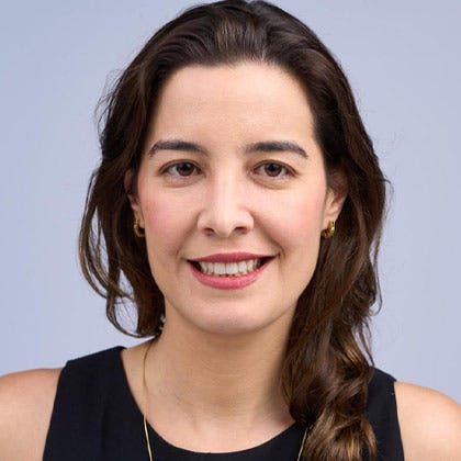 Uma mulher com cabelos castanhos longos e olhos escuros. Ela usa uma regata preta e está sorrindo