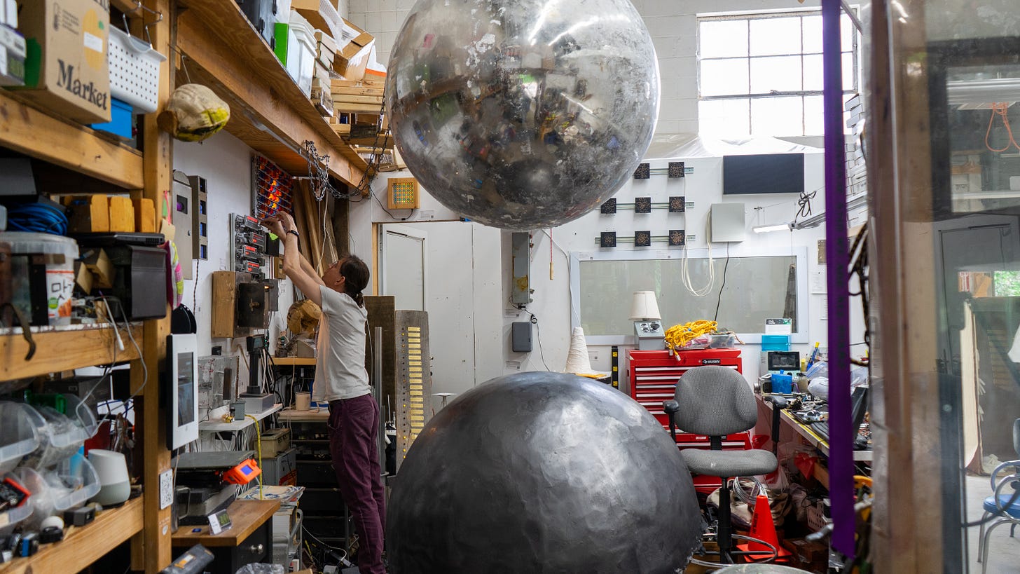The metallic steel ball from the installation hangs at the center of the image, and the center of Combs’ studio.  Below it is a copy in black that Combs is still forming. Combs can be seen on the left side, reaching up to adjust one of the many machines on the wall that light and blink. The studio looks messy to the untrained eye, tools and materials seem to be everywhere, but an artist knows there is a precise logic formed by habit, and everything is actually in its rightful place. 
