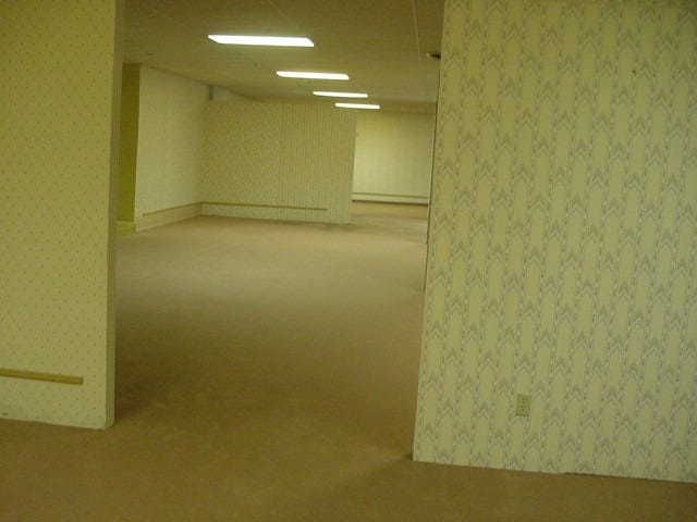 An empty maze of yellow-walled rooms lit by fluorescent ceiling panels. Beige carpeting, repeating wallpaper, and a lack of furniture or landmarks make the abandoned space feel strangely familiar yet deeply unsettling. An empty maze of yellow-walled rooms lit by fluorescent ceiling panels. Beige carpeting, repeating wallpaper, and a lack of furniture or landmarks make the abandoned space feel strangely familiar yet deeply unsettling.