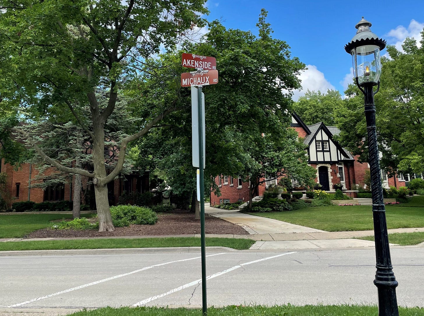In the foreground, Akenside Road and Michaux Road street signs beside a gas street light. Mature trees and Tudor-style house are in the background.  