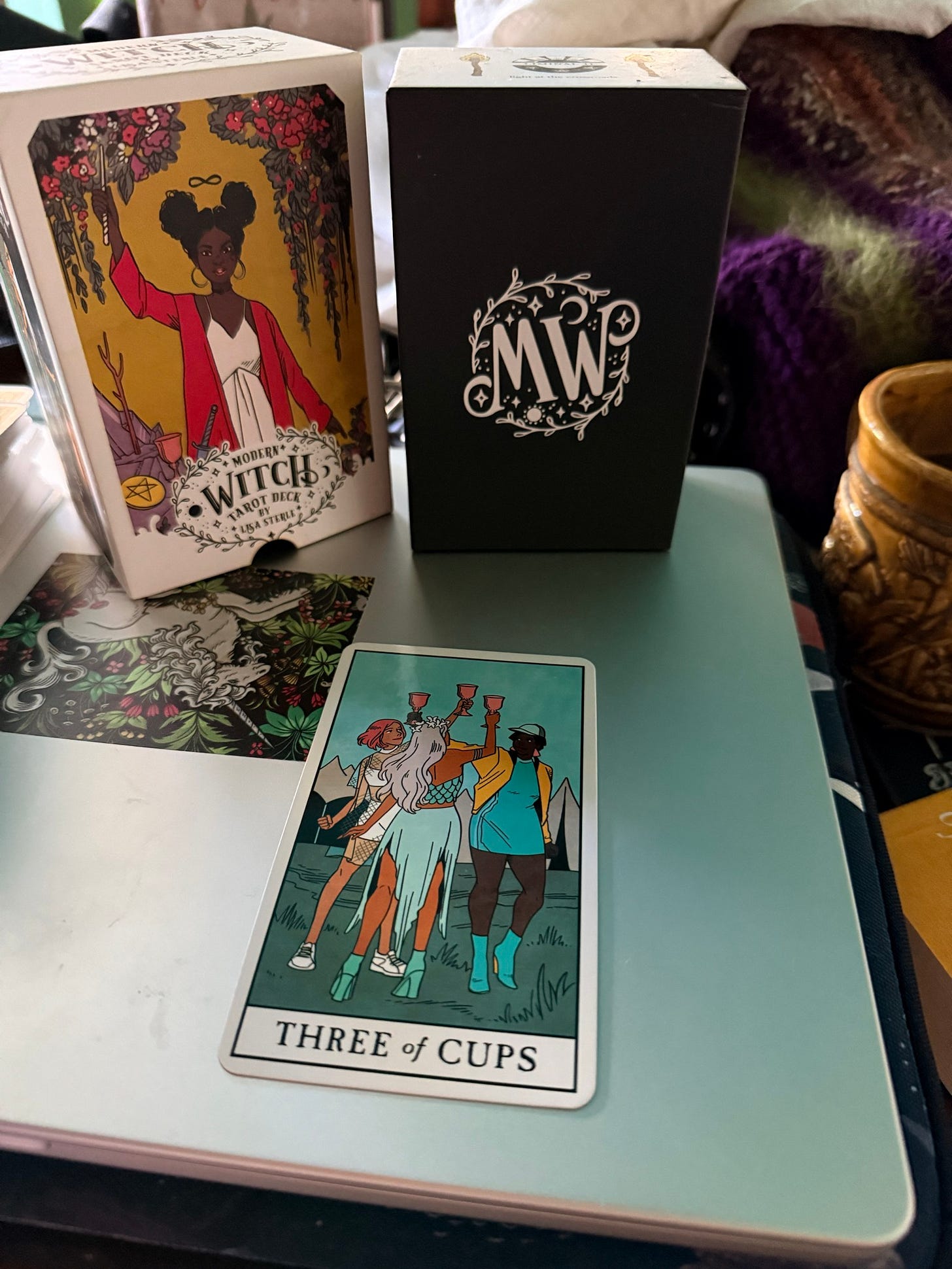 The Modern Witch Tarot deck packaging by Lisa Sterle, a black MWT box and the Three of Cups tarot card displayed together on a pale green surface. The Modern Witch Tarot deck packaging by Lisa Sterle, a black MWT box and the Three of Cups tarot card displayed together on a pale green surface.