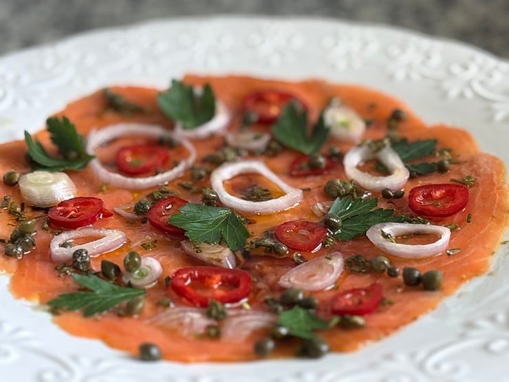 Smoked salmon carpaccio elegantly presented with capers, red chili slices, shallots, and fresh parsley on an intricate white plate.