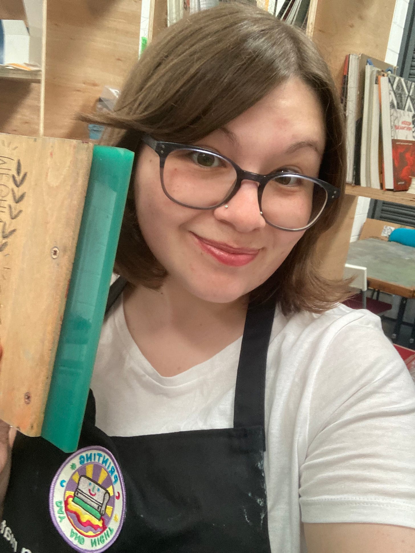 Picture of Megan, a woman in an art studio wearing dark framed glasses, with brown hair, a white T-shirt and a black apron.