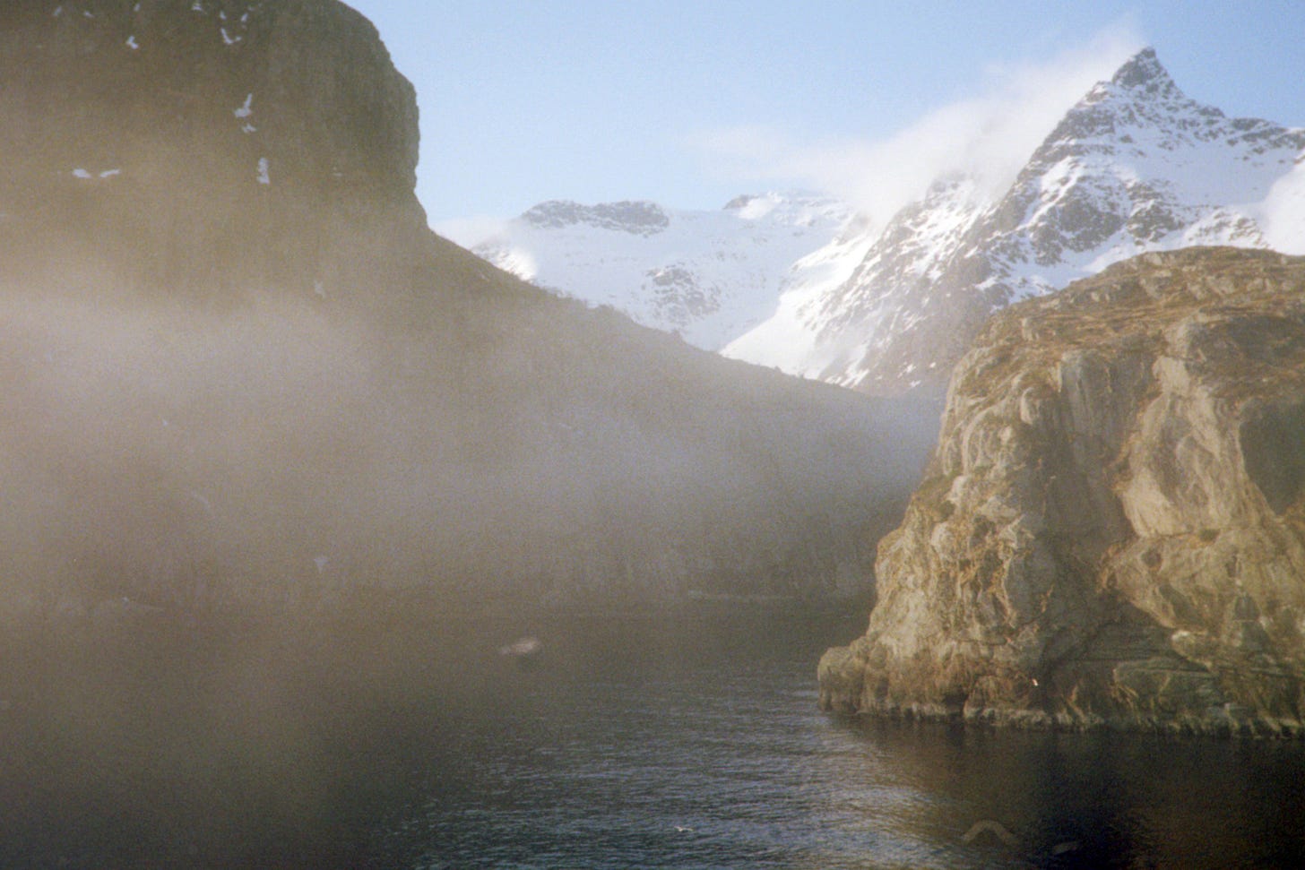 a dreamy, misty mountain seaside, the water is black and a snow-capped mountain rises behind a moss-covered cliffside
