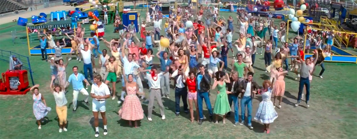 Crowd of excited school kids wave from the ground as Sandy and Danny ascend into the sky in Greased Lightning - still from 1978 film Grease