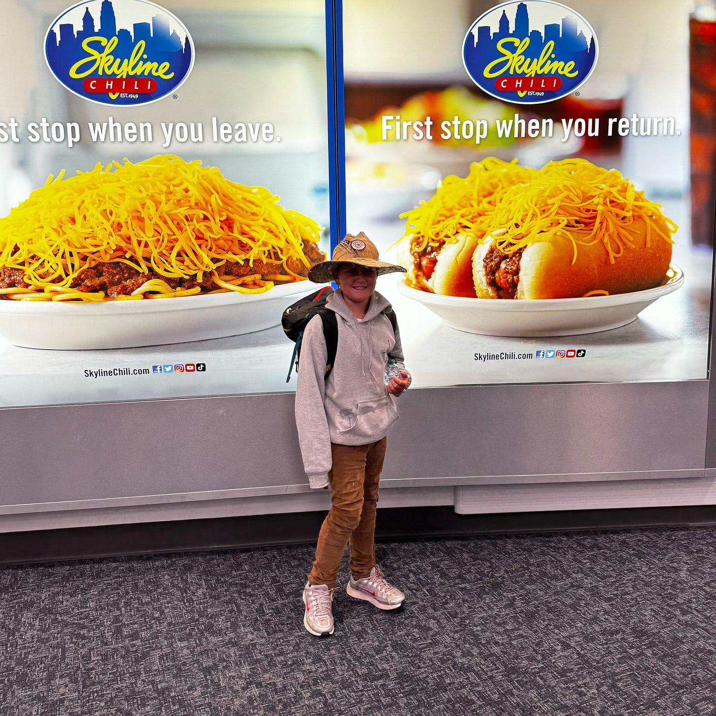 Capture the joy of travel and family moments in this candid image of Andrew's son at the airport. With a casual look, backpack, and a bright backdrop featuring Skyline Chili, this picture perfectly reflects the excitement of returning home and the spirit of new adventures. Ideal for stories on family journeys, memorable trips, and the special bond between parent and child during life’s transitions.
