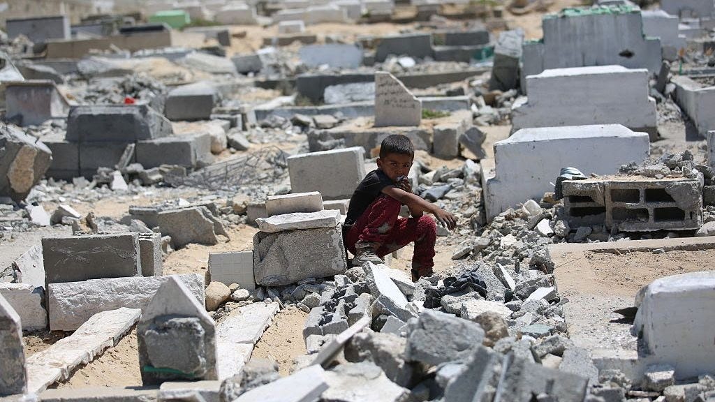 A child with black hair and brown eyes, who is wearing a blue tshirt and red trousers, sits among white and grey rubble.