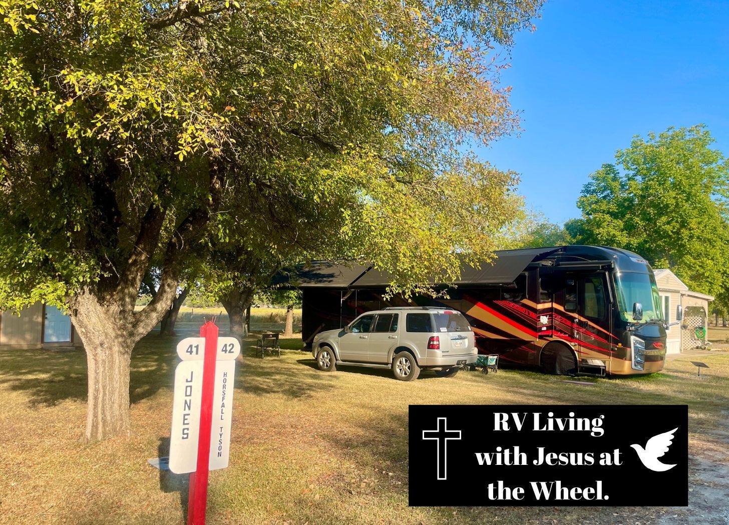 Big Red, our black and red Entegra Anthem motorhome, parked beneath shady trees at our RVICS Village home base in Smithville, Texas, with our site marker and RV Living with Jesus at the Wheel logo in the foreground on a peaceful sunny afternoon.