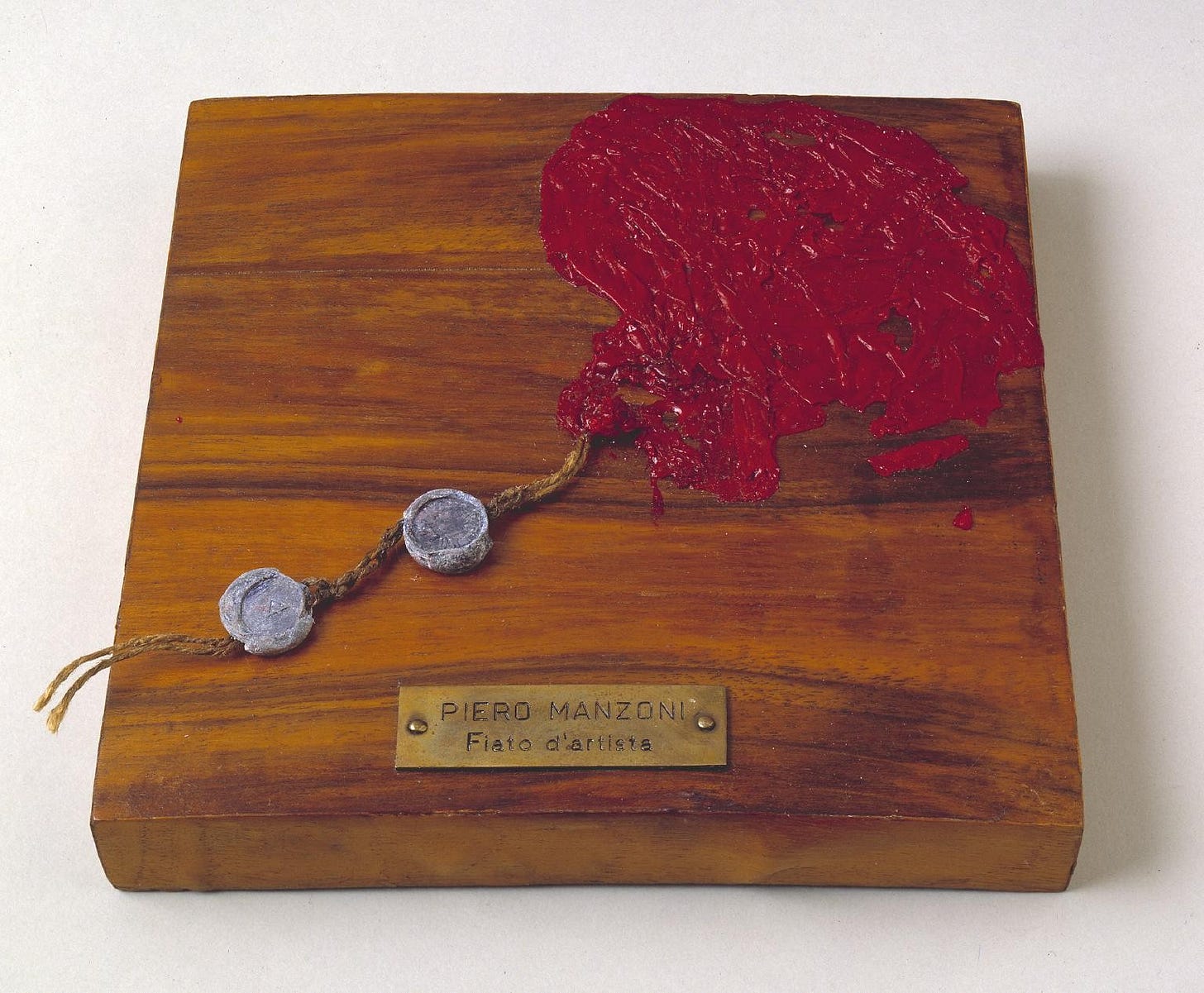 Piero Manzoni’s work Artist’s Breath: the dried remains of a red balloon tethered to a rectangular block of wood. It is labelled Piero Manzoni, Fiato d’Artista