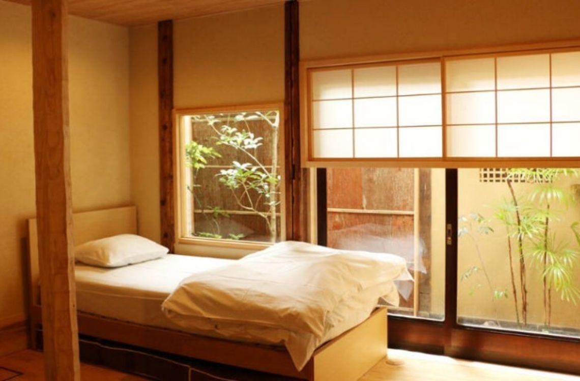 A simple, light-filled bedroom at Hibari Hostel with a single bed, shoji sliding doors, and a window overlooking a small garden with bamboo and greenery.