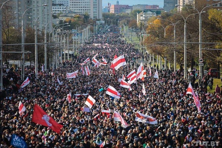 This may contain: a large group of people holding flags in the middle of a street with trees and buildings behind them This may contain: a large group of people holding flags in the middle of a street with trees and buildings behind them