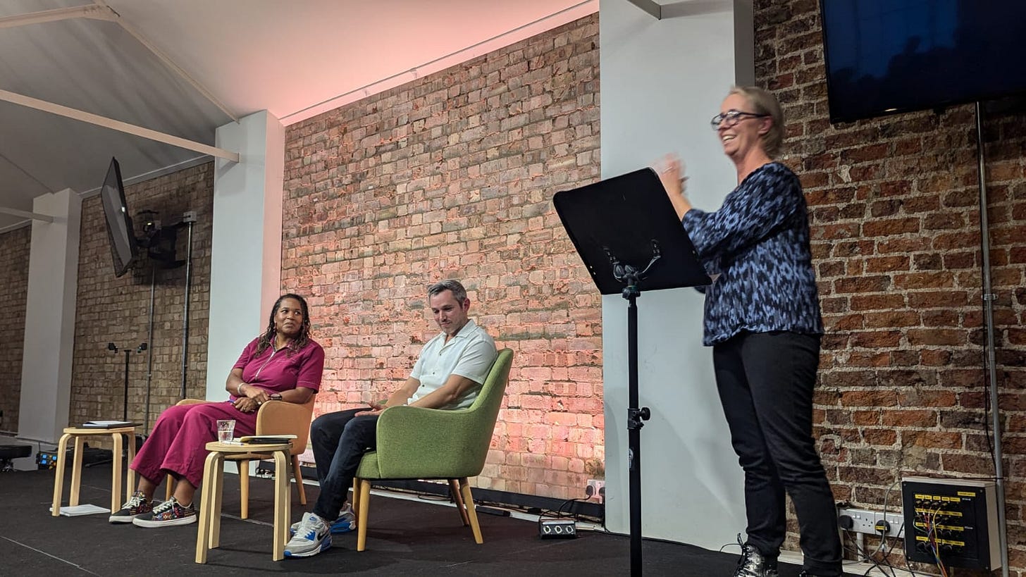A women is standing up at a lectern smiling while she talks to an unseen audience. There is a brick wall behind her. To her left is Jake, sitting in a chair in a nice white shirt. To his left is a black women with long hair and a red/purple jump suit sitting next to him listening to the speaker. 