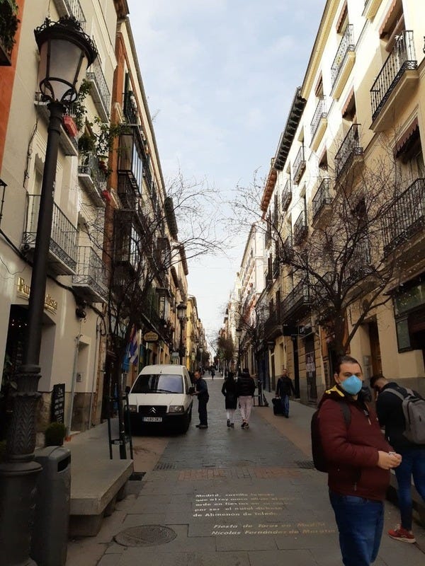 Bairro de Las Letras, em Madrid. Foto de Edmara Galvão. Descrição da Imagem: Uma rua estreita, com um escrito em dourado no chão. Na foto, um carro branco e diversas pessoas passeiam pela rua ladeada por árvores e prédios de três andares. No lado esquerdo, em primeiro plano, uma luminária. No lado direito, um homem de blusa vinho, calça jeans e máscara azul-claro olha para a câmera. Ele tem cabelo preto preso para trás e é branco.Uma rua estreita, com um escrito em dourado no chão. Na foto, um carro branco e diversas pessoas passeiam pela rua ladeada por árvores e prédios de três andares. No lado esquerdo, em primeiro plano, uma luminária. No lado direito, um homem de blusa vinho, calça jeans e máscara azul-claro olha para a câmera. Ele tem cabelo preto preso para trás e é branco.