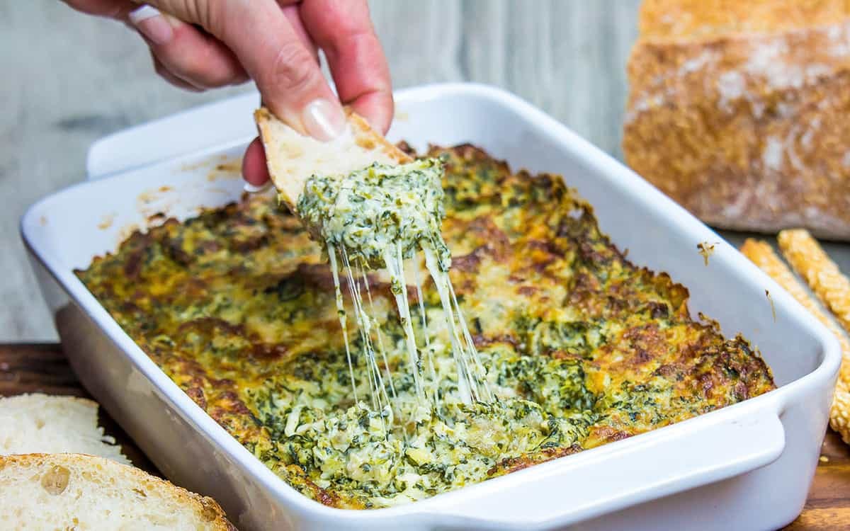 Warm baked spinach dip with melted cheese being scooped from a baking dish with a piece of bread.