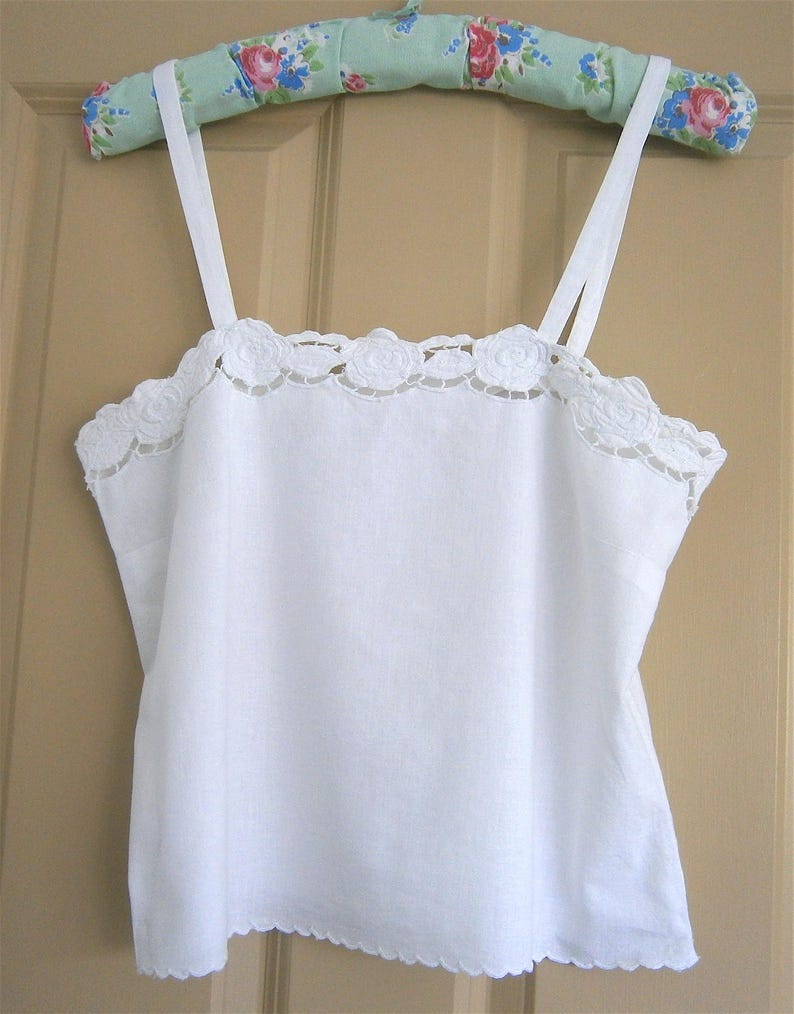 May include: A white camisole with delicate lace trim along the neckline and straps. The camisole is hanging on a floral-patterned hanger. The bottom hem has a scalloped edge.