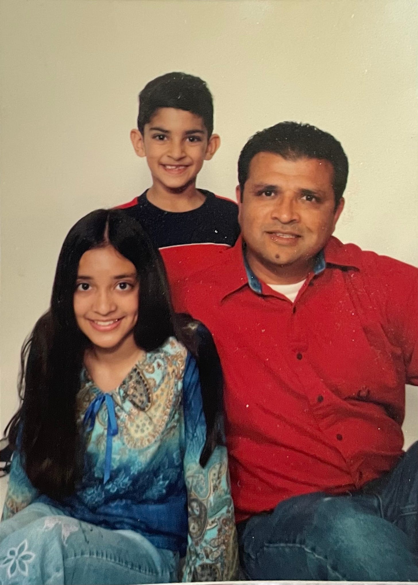 A preteen wears a paisley top and jeans with flower designs. She has long black hair and smiles. A young boy stands behind her, with a red and black shirt, smiling. A man in his forties sits in front of the boy and next to the girl, wearing a red shirt and smiling.