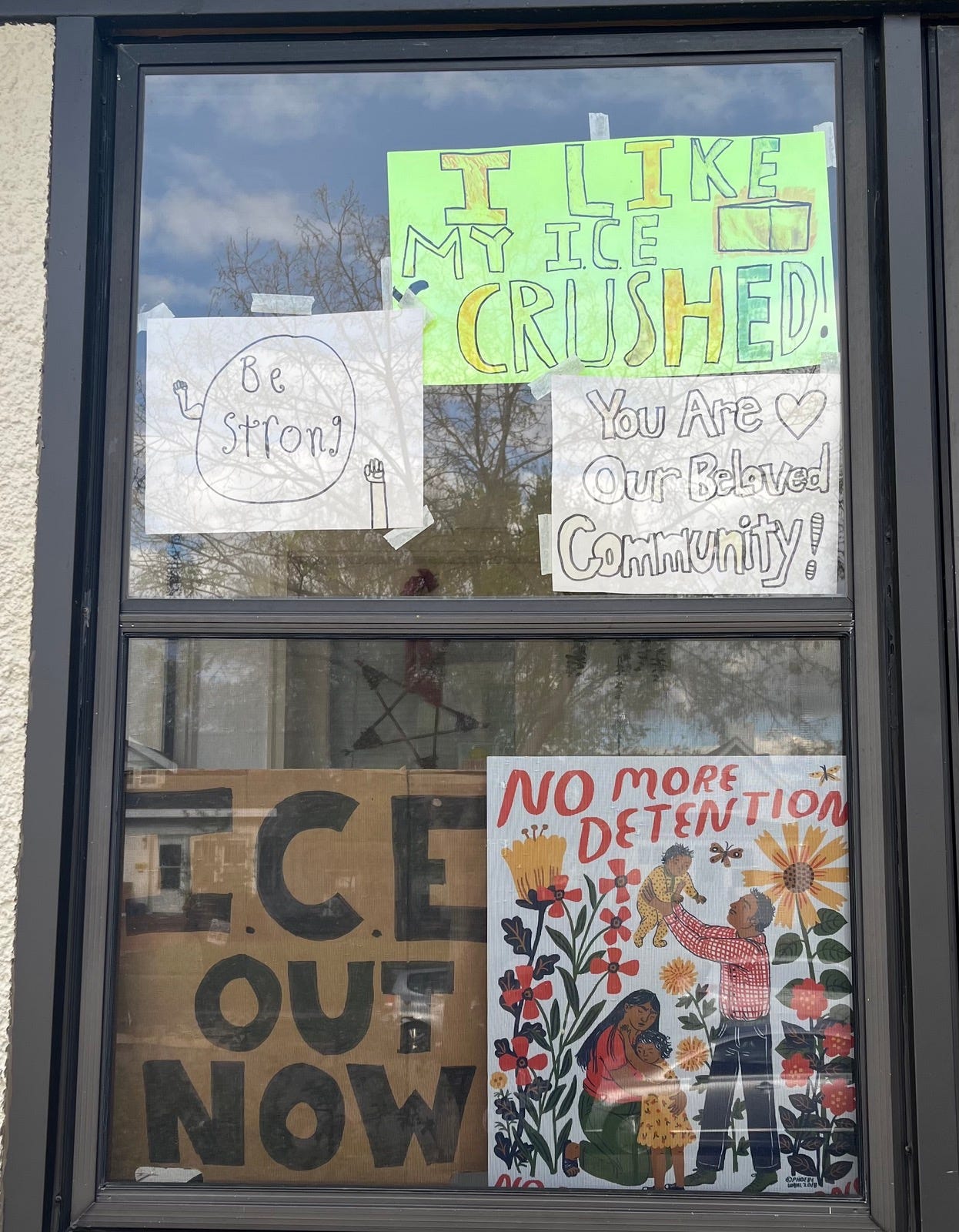 Window covered with handmade signs and posters advocating for immigrant rights and community support. Messages include “I like my ICE crushed,” “Be strong,” “You are our beloved community,” “No more detention,” and a large “ICE out now” sign, alongside an illustrated poster of a family surrounded by flowers.