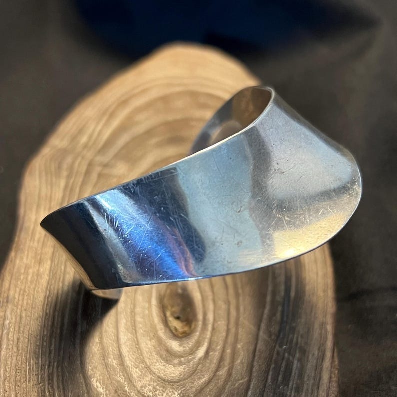 May include: A polished silver cuff bracelet with a curved, open design. The bracelet has a smooth, reflective surface and is displayed on a wooden surface with a natural grain pattern. The cuff is a modern piece of jewelry.
