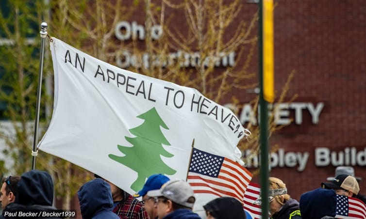 A white flag bearing the words 'An Appeal to Heaven,' featuring a green pine tree, with the American flag displayed beneath it. A white flag bearing the words 'An Appeal to Heaven,' featuring a green pine tree, with the American flag displayed beneath it.