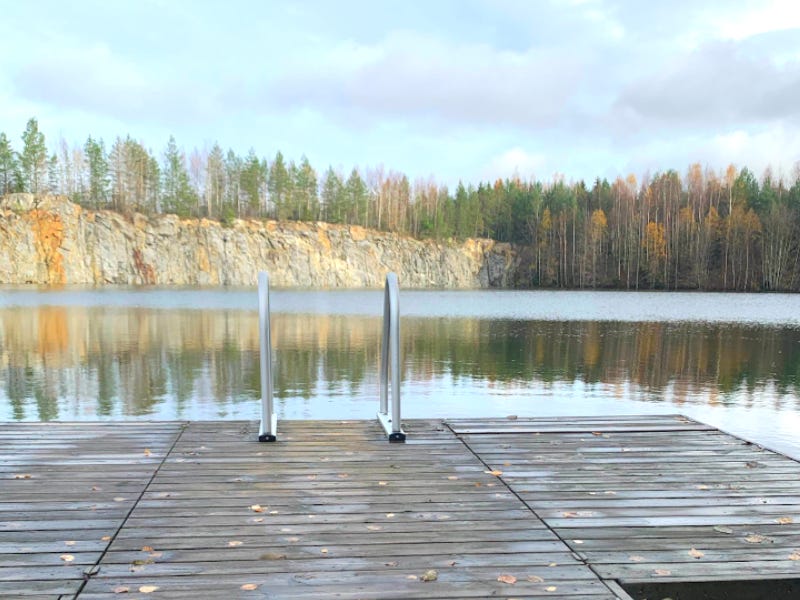 A small dock with two metal railings into the water is in the foreground. Ahead is clear water, a rock ridge and birch and pine trees.