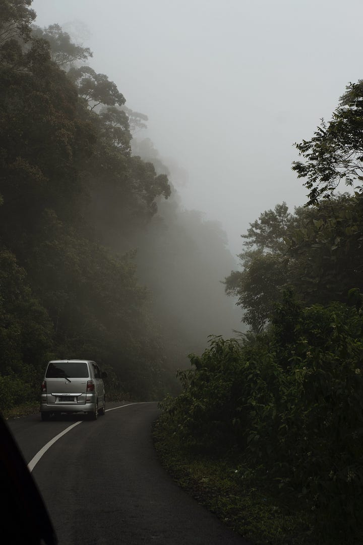 A van crosses a strech of the jungle through the fog. A hand says goodbye to a schoolbus with kids siting on the roof of their bus. In the background is the active volcano Mount Sinabung