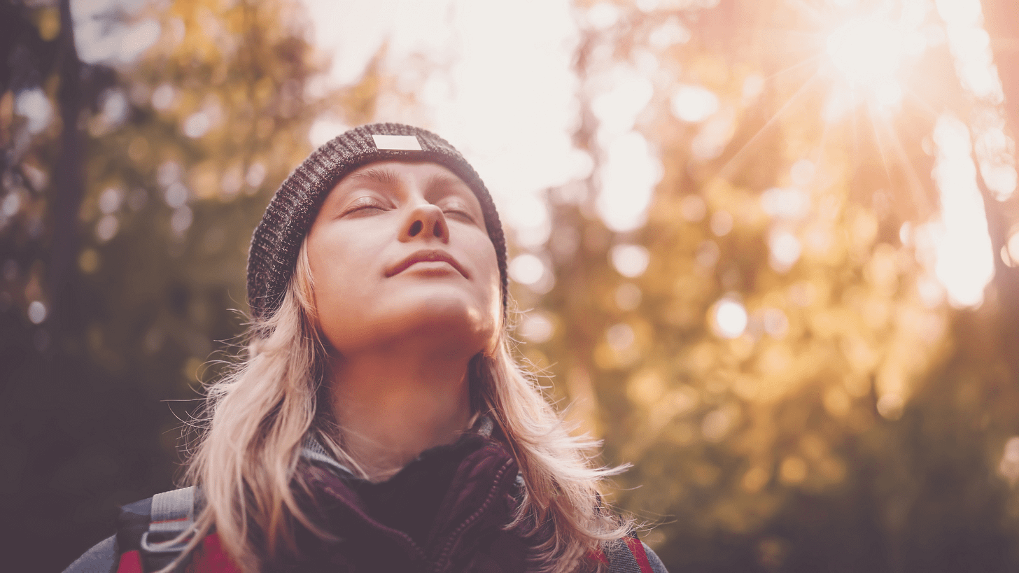 Portrait of a woman outdoors with her eyes closed, wearing a knit hat and scarf, bathed in soft sunlight filtering through the trees. She appears calm and reflective, surrounded by a natural, golden-hued forest setting
