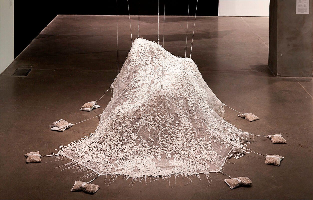 A delicate, sculptural installation of white lace fabric draped like a shrouded figure in the centre of a gallery floor. The lace is densely embroidered with small white floral or petal-like elements. The fabric is anchored at multiple points by cords extending to small sandbags arranged around the perimeter. The piece is softly lit, creating shadows and texture against the smooth concrete floor.