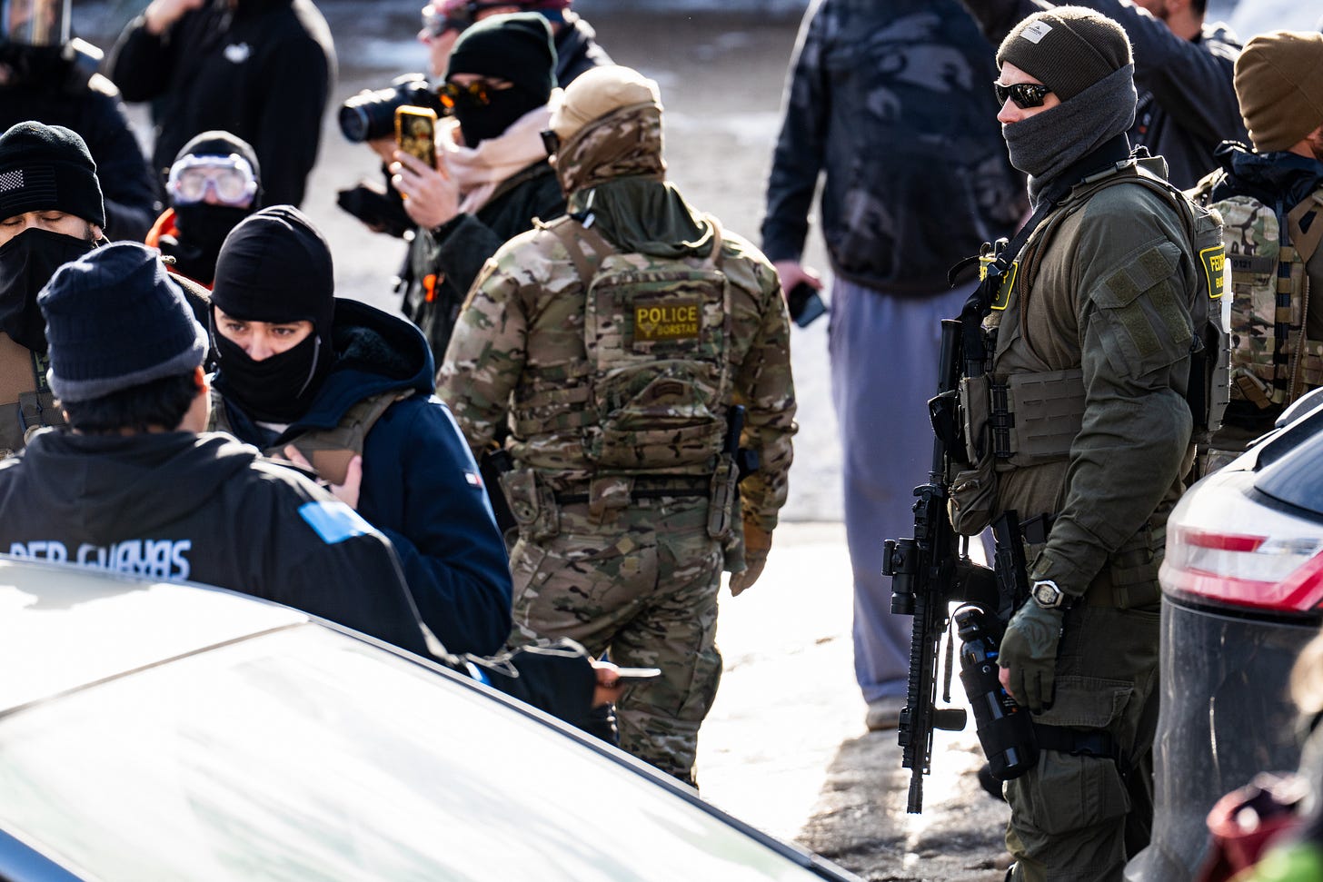 Several armed, masked men stand facing a crowd of people holding cell phones,  while a masked man in the center stands in an aggressive posture toward a man who is leaning up against a car. Original source description: "Federal agents ram a man's vehicle and demand identification at Park Avenue and 35th Street in Minneapolis on January 12, 2026. The Latino man says he was let go once they realized he was a US citizen. While doing so, a crowd as well as more officers continued to arrive before releasing tear gas and pepper spraying members of the media and their cameras.  Park and 35th Street is 2 blocks away from where ICE agents shot and killed Renee Good on January 7, 2026."