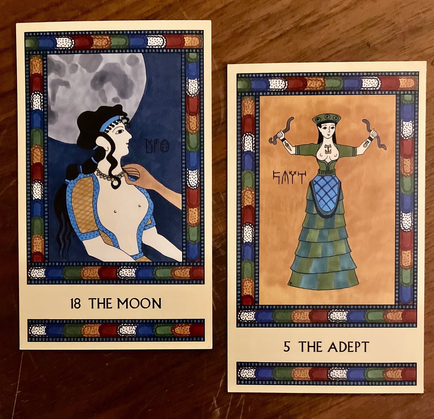 Two Minoan Tarot cards side by side on a brown wood surface. Both cards have multicolored borders. The Moon card, on the left, shows a Minoan woman from the waist up, seated and facing right, while a man's hand adjusts her necklace. The Full Moon looms large behind her head in a dark sky. The Adept card, on the right, shows the Minoan Snake Goddess in a long tiered skirt, holding up a snake in each hand. The background around her is deep gold.