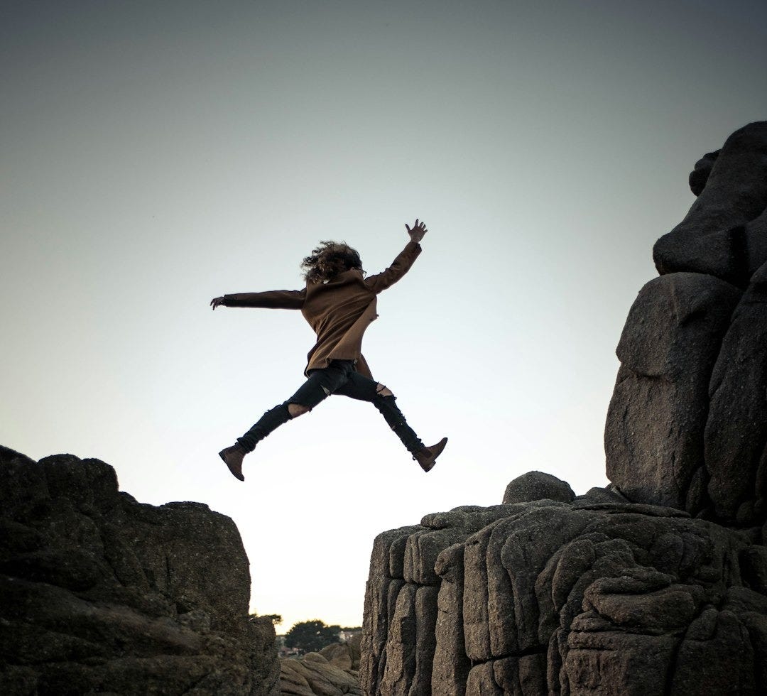 person jumping on big rock under gray and white sky during daytime