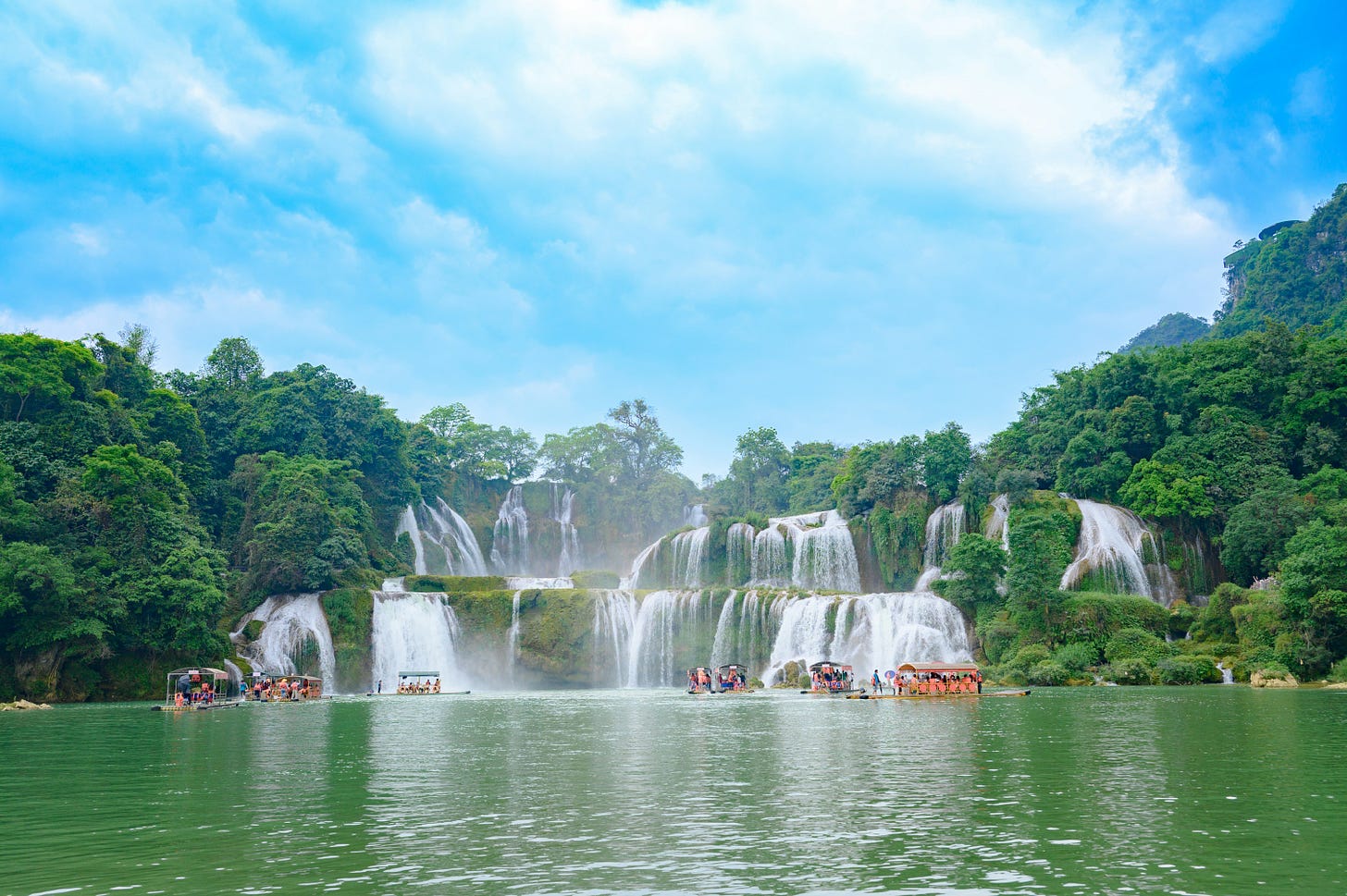 wide, panoramic waterfall cascading across multiple lush green tiers, with a bamboo raft floating on the river at its base.