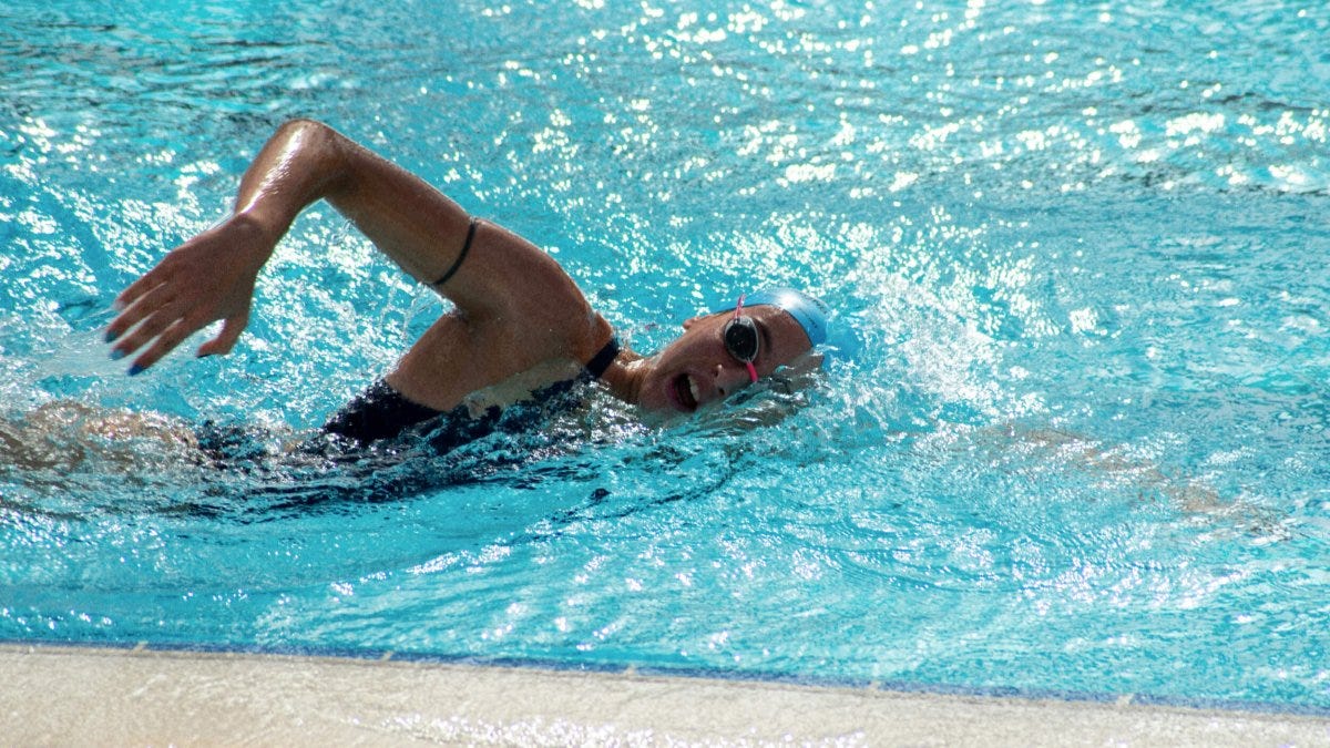 A swimmer wearing goggles and a swim cap performs the freestyle stroke in an outdoor pool near the pool edge.